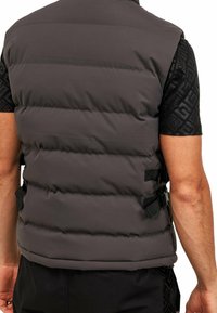 Gilet imbottito grigio scuro con cuciture orizzontali, strap regolabili sui lati e una superficie liscia. Indossato sopra una camicia a maniche corte con fantasia.