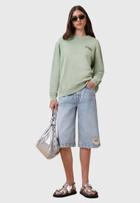 Felpa verde chiaro con una vestibilità casual, abbinata a pantaloncini in denim chiaro sfiniti. Il modello porta una borsa metallica argento e indossa sandali chunky.