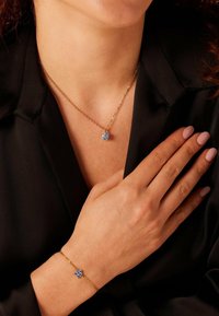 Vrouw in een zwarte blazer, met een gouden ketting en armband, beide met een blauwe edelsteen bloemenhanger, hand rustend op de borst.