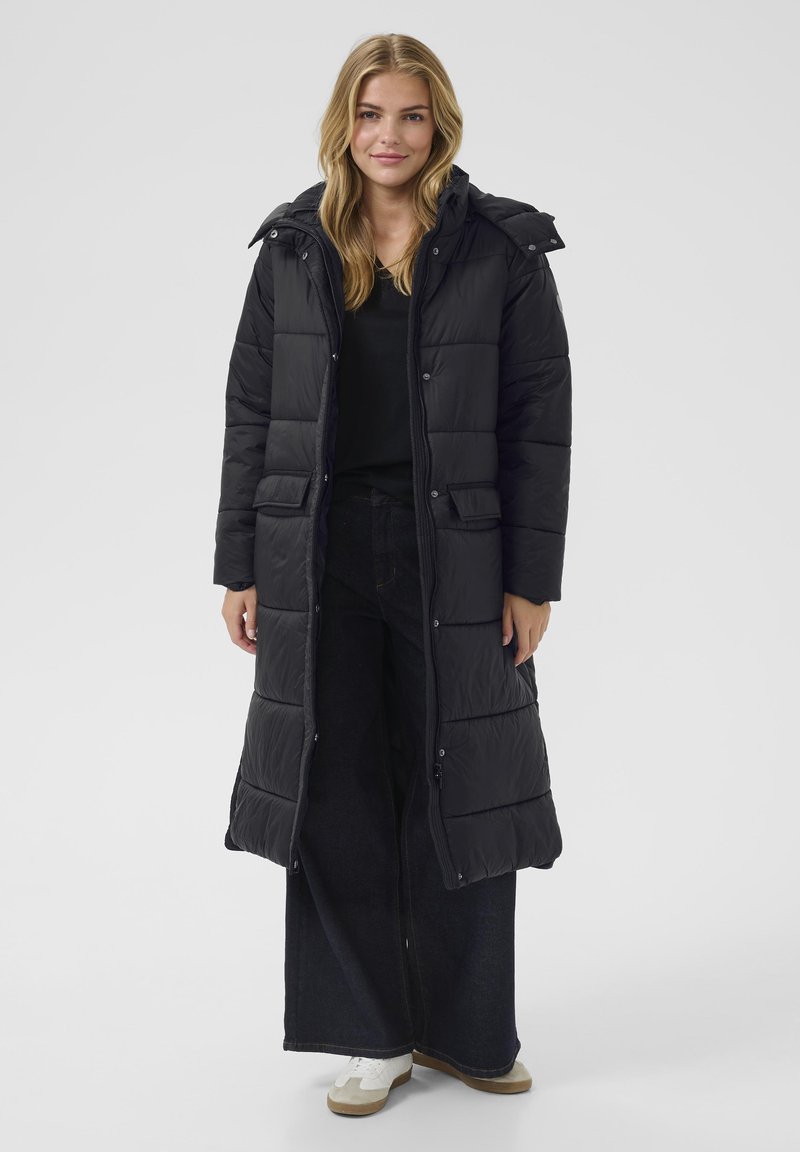 Abrigo acolchado negro largo con capucha, que presenta secciones acolchadas, bolsillos delanteros y un zipper oculto. Lleva puesto sobre pantalones de mezclilla oscuros.