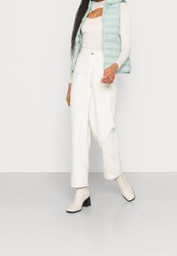 Woman wearing cream long-sleeve top, light blue quilted vest, high-waisted white pants, and cream heeled ankle boots, standing on a gray floor.