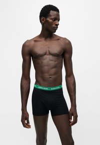 Homme musclé à la peau foncée portant un boxer noir avec une ceinture verte portant l'inscription Björn Borg, debout et regardant vers sa gauche.