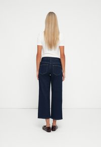 Jeanși largi de culoare albastru închis cu talie înaltă, textură de denim și detalii subtile de cusătură. Asortați cu pantofi băieți maro și un top alb.