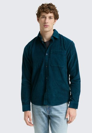 Chemise en velours côtelé teal avec un devant boutonné, des manches longues et une poche poitrine, associée à un jean bleu clair. Surface en tissu texturé.