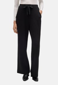 Pantalones de pierna ancha negros con una textura suave, con cintura ajustable y bolsillos laterales. Combinados con una blusa ajustada de manga larga y zapatos negros.