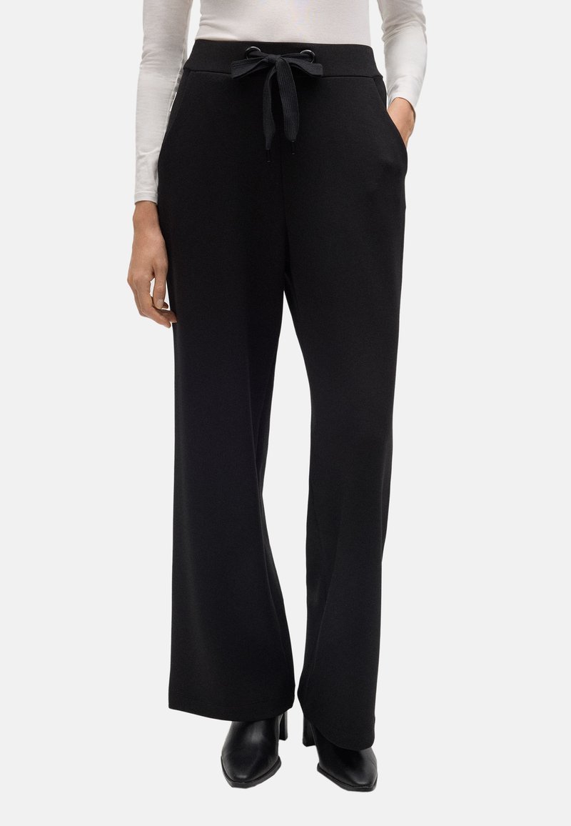 Pantalones de pierna ancha negros con una textura suave, con cintura ajustable y bolsillos laterales. Combinados con una blusa ajustada de manga larga y zapatos negros.