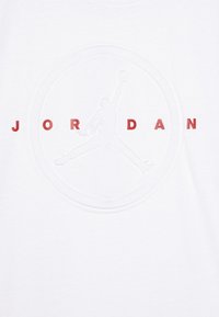 T-shirt bianco in cotone con una grafica in rilievo di una figura che salta e il testo "JORDAN" in rosso, centrato sul petto.