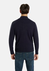 Tumši jūras zilas adītas džemperis ar ribotu apkakli un aprocēm, ar garām piedurknēm un pieguļošu dizainu. Valkāts virs ziliem džinsiem.