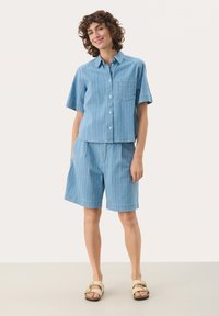 Vaaleansininen raidallinen lyhythihainen napitettu paita, jossa on rintatasku, yhdistettynä saman sävyisiin sinisiin shortseihin. Asusteena beige sandaalit.