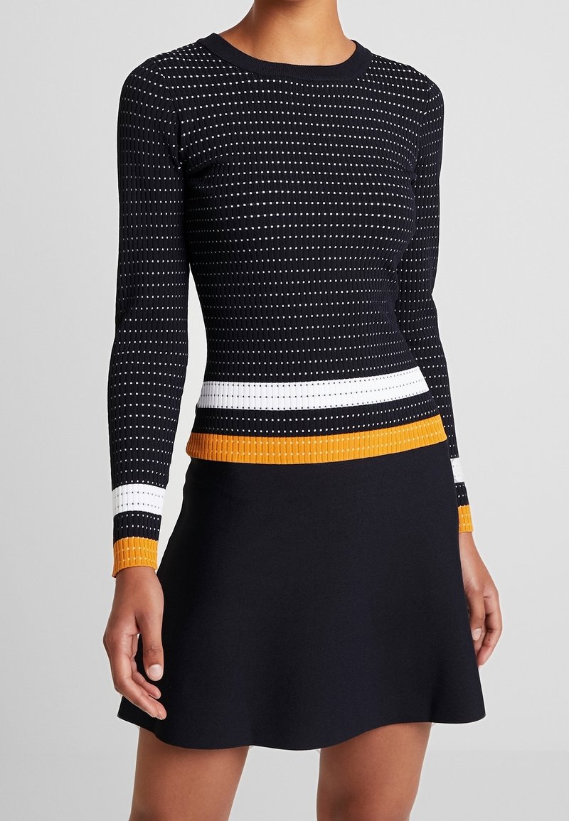 Donna che indossa un maglione nero a maniche lunghe attillato con strisce bianche e arancioni e una gonna nera su uno sfondo semplice.