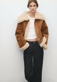 Bruine suède jas met een pluizige beige kraag en manchetten, ritssluiting, gecombineerd met donkerblauwe jeans met hoge taille. Effen wit topje eronder.