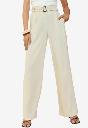 Pantaloni beige a gamba larga con vita regolabile, caratterizzati da pieghe frontali, tasche laterali e un tessuto strutturato, abbinati a sandali neri con cinturini.