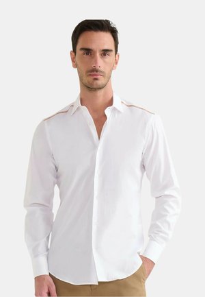 Uomo che indossa una camicia bianca a maniche lunghe con bottoni e cuciture color sabbia sulle spalle, abbinata a pantaloni beige, in piedi con una mano in tasca.