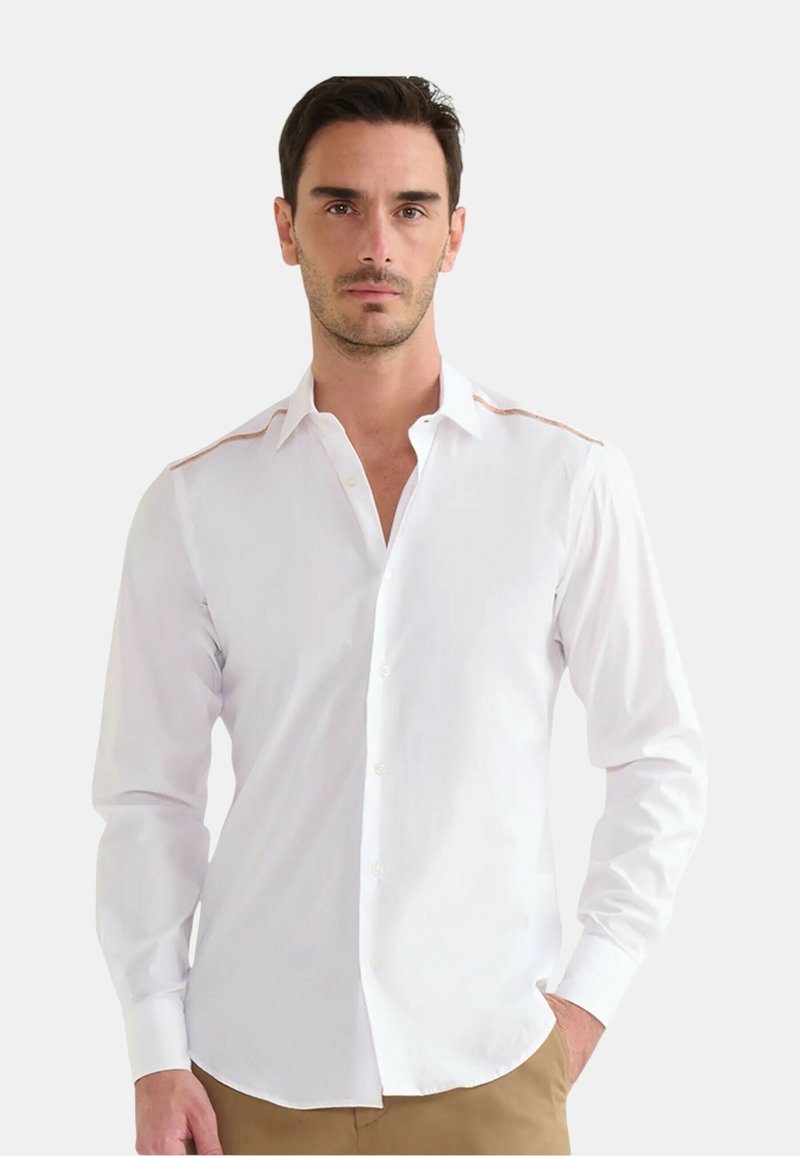 Uomo che indossa una camicia bianca a maniche lunghe con bottoni e cuciture color sabbia sulle spalle, abbinata a pantaloni beige, in piedi con una mano in tasca.