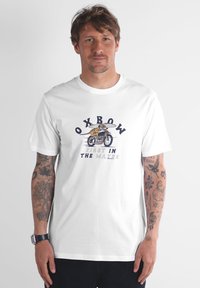T-shirt en coton blanc avec un motif représentant "Oxbow" et un surfeur à vélo, texte indiquant "FIRST IN THE WATER". Manches courtes, col rond.