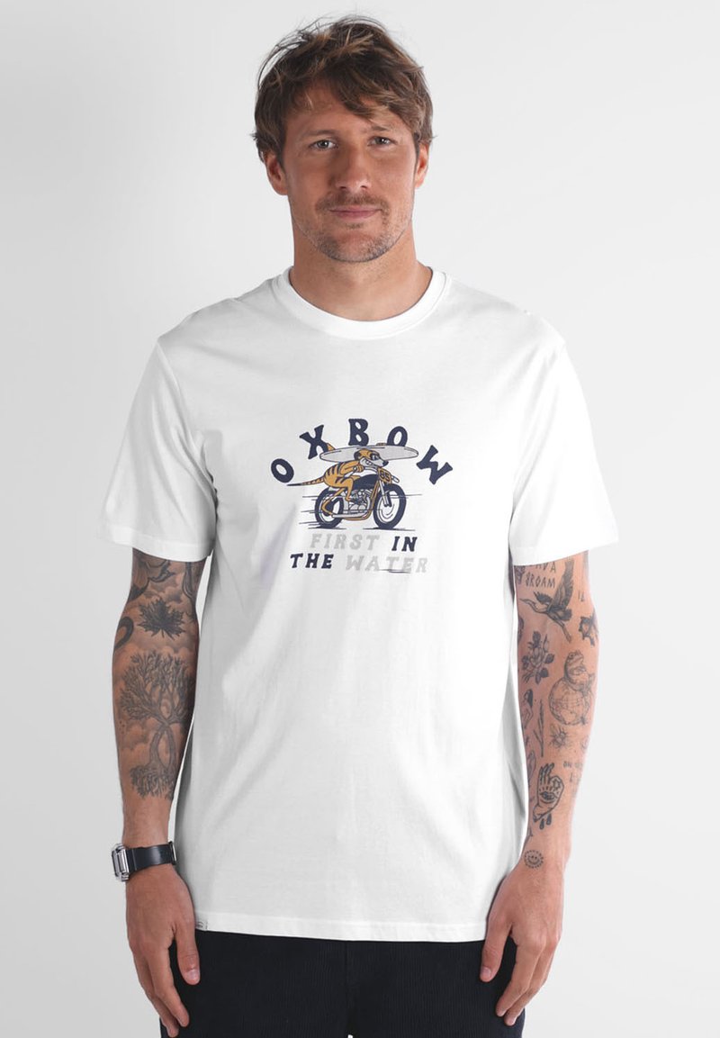 T-shirt en coton blanc avec un motif représentant "Oxbow" et un surfeur à vélo, texte indiquant "FIRST IN THE WATER". Manches courtes, col rond.