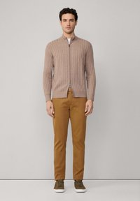 Beżowy kardigan z dzianiny w splot kablowy z zamkiem z przodu, połączony z brązowymi chinosami i zielonymi trampkami. Materiał jest teksturowany i gładki.