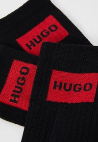 Calcetines negros con un panel rectangular rojo en el que está tejido en negro la palabra "HUGO". La textura acanalada y los colores contrastantes mejoran la visibilidad.