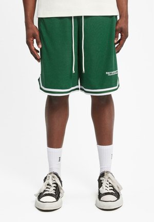 Grüne Sportshorts mit weißen Akzenten und Kordelzug, ausgestattet mit einem strukturierten Stoff. Getragen mit weißen gerippten Socken und schwarzen Turnschuhen.
