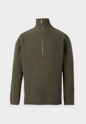 Homme portant un pull kaki à demi-fermeture éclair, un tee-shirt blanc, un jean bleu et des chaussures noires, debout avec les mains dans les poches devant un fond uni.