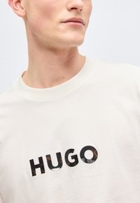 Νεαρό άτομο που φοράει λευκό μπλουζάκι με στρογγυλή λαιμόκοψη και μαύρα, διακοσμητικά γράμματα που σχηματίζουν τη λέξη "HUGO" στο στήθος.