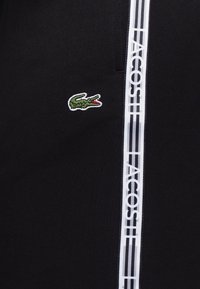 Svart sweatshirt med en liten grön krokodil-logotyp och vit vertikal "LACOSTE"-branding på sidan. Slät textur, avslappnad design.