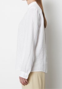 Chemise en lin blanc à manches longues, col mao et poignets à boutons. Le tissu a une finition texturée. Associée à un pantalon beige clair.