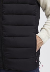 Svart puffervest med horisontal quilt, med glidelås lommer og kontrasterende røde og hvite glidelåsgrep. Myk strikket gensermansjett synlig.