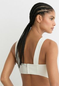 Valkoinen säädettävä rintaliivi, jossa on leveät olkaimet, kuvioitu kangas ja takana vetoketju. Siinä on sileä, minimalistinen muotoilu.