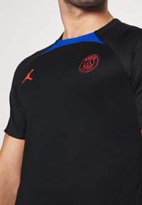 Czarna koszulka sportowa z niebieskimi akcentami, z czerwonym logo Paris Saint-Germain oraz czerwonym emblematem Jumpman na klatce piersiowej.