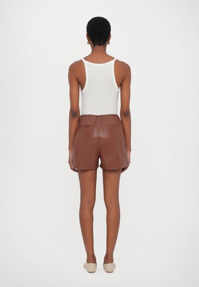 Brune lædershorts med en afslappet pasform, som har en elastisk talje og to baglommer, sammen med en hvid tanktop.