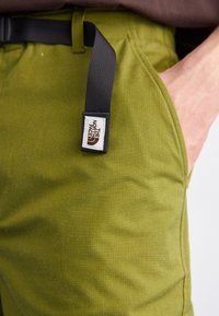 Olive zelene hlače z teksturirano tkanino, opremljene z črnimi nastavljivimi pasom in belim oznako s perlaste barve. Vidna stranska žep.