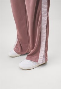 Světlé růžové tepláky se lesklými bílými bočními pruhy, v kombinaci s bílými teniskami s texturovanou podrážkou. Tepláky mají volný střih a měkkou látku.