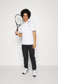 Vit polo t-shirt med svart kant, svarta träningsbyxor med vita sidostreck och vita sneakers. Håller en svart tennisracket.