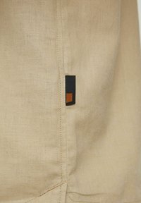 Beige Stoff mit einer weichen Textur und subtilen Webdetails. Besitzt ein kleines, rechteckiges schwarz-oranges Etikett in der Nähe der Naht.
