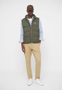 Maslinasto zeleni pufasti prsluk s patentnim zatvaračem, nosi se preko svijetloplavog košulje. Uparen s bež cargo hlačama i bijelim tenisicama. Uključena smeđa kapa.