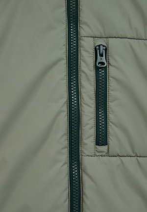 Giacca a vento verde chiaro realizzata in tessuto con cerniere verde scuro. Il materiale presenta una texture liscia con imbottitura trapuntata e dettagli delle cerniere in contrasto.