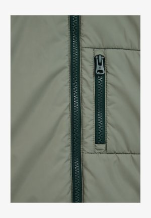 Vaalea vihreä puhvitakki, jossa on tumma vihreä vetoketju. Materiaali on sileä ja siinä on tikattu täyte sekä kontrastiväriset vetoketjut.