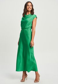Long green satin dress with a draped neckline and gathered waist. Features short cap sleeves and a flowing hemline. Worn with gold strappy heels.