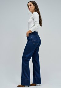 Mujer con cabello largo vestida con una blusa blanca de manga larga, jeans de tiro alto de pierna ancha en azul oscuro y tacones puntiagudos marrones, de pie de lado.
