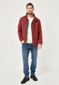 Red synthetic jacket with gray lining, front zip, two chest pockets, worn over a white sweatshirt, paired with blue jeans and black shoes.