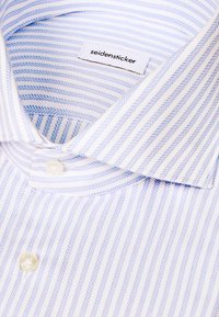 Gros plan d'une chemise blanche pliée avec des rayures en chevrons bleu clair et une étiquette "seidensticker" visible à l'intérieur du col.