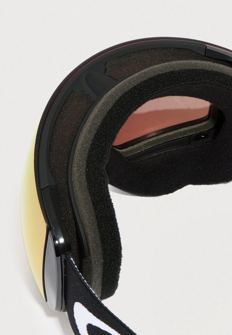 Lunettes de ski avec verres teintés jaunes, épais rembourrage en mousse noire et sangle noire réglable avec logo blanc sur fond blanc.