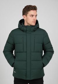 Grüne Steppjacke mit Kapuze, die gesteppten Abschnitte, zwei Brusttaschen und Kordelzugverstellungen aufweist. Das Material wirkt glatt und strapazierfähig.