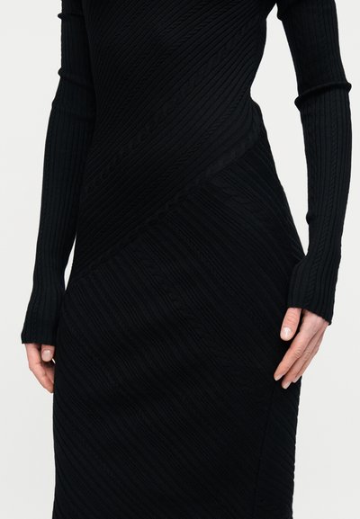 Μαύρο πλεκτό φόρεμα με ανάγλυφα διαγώνια σχέδια, μακριά μανίκια και μανσέτες με άνοιγμα για τον αντίχειρα, εμφανιζόμενο σε ένα άτομο από τη μέση έως τα χέρια.