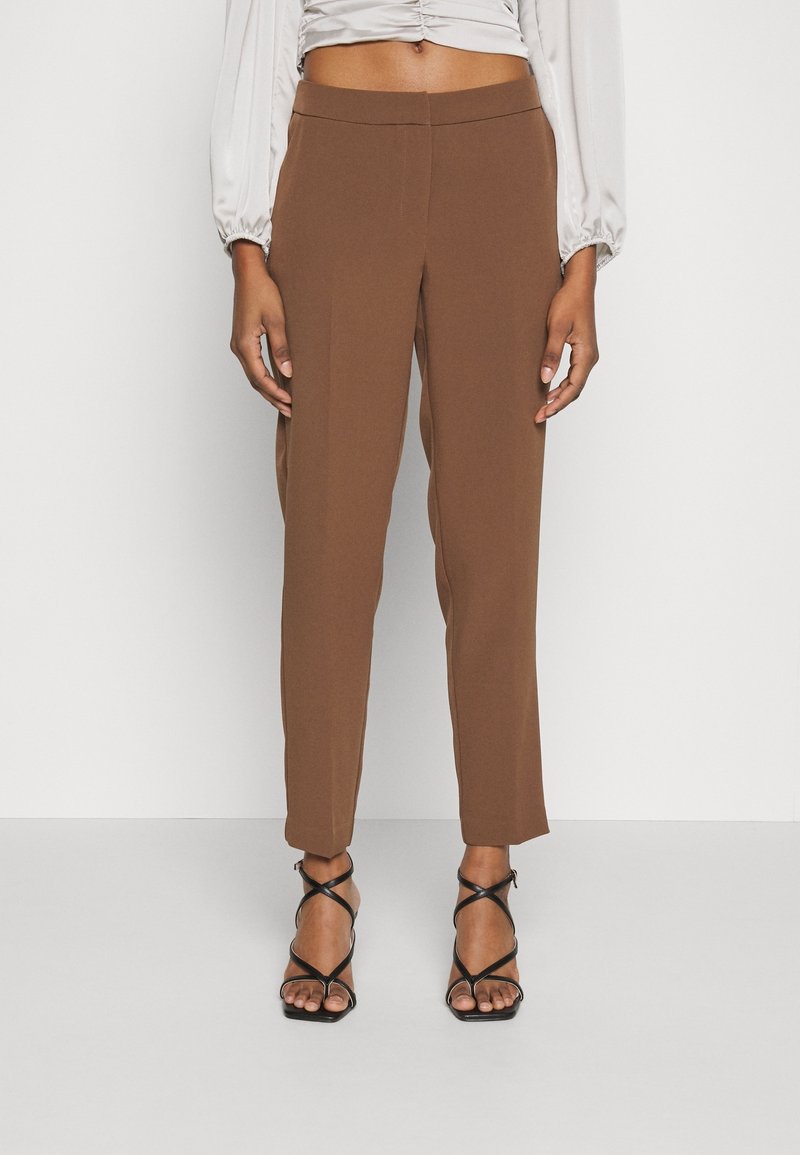 Personne portant un pantalon marron taille haute et fuselé, avec un chemisier beige froncé et des sandales à talons avec lanières noires, debout devant un fond uni.