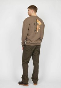 Sudadera marrón con un gráfico de un oso en la parte posterior, combinada con pantalones verde oscuro y zapatos marrones. Tejido suave, corte relajado, puños acanalados.