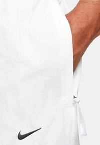 Vit sportjacka i lättviktstyg, med en svart Nike-logotyp och dragsko-detaljer på sidorna. Slät textur.