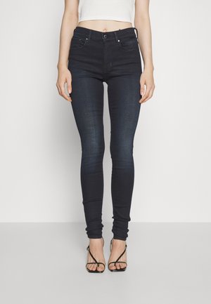 Jeans oscuros ajustados de cintura alta, con diseño de cinco bolsillos y bucles para cinturón. Llevados con tacones negros de tiras. Tejido de mezclilla suave y elástico.