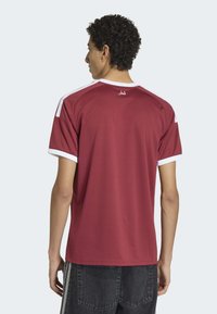 Vinröd sporttröja med korta ärmar, vita detaljer vid kragen och ärmarna samt ett texturerat tyg. Arabisk text visas på baksidan.